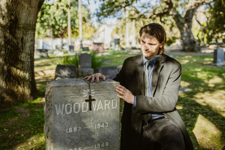 A man kneels in a cemetery, reflecting at a gravestone under daylight.